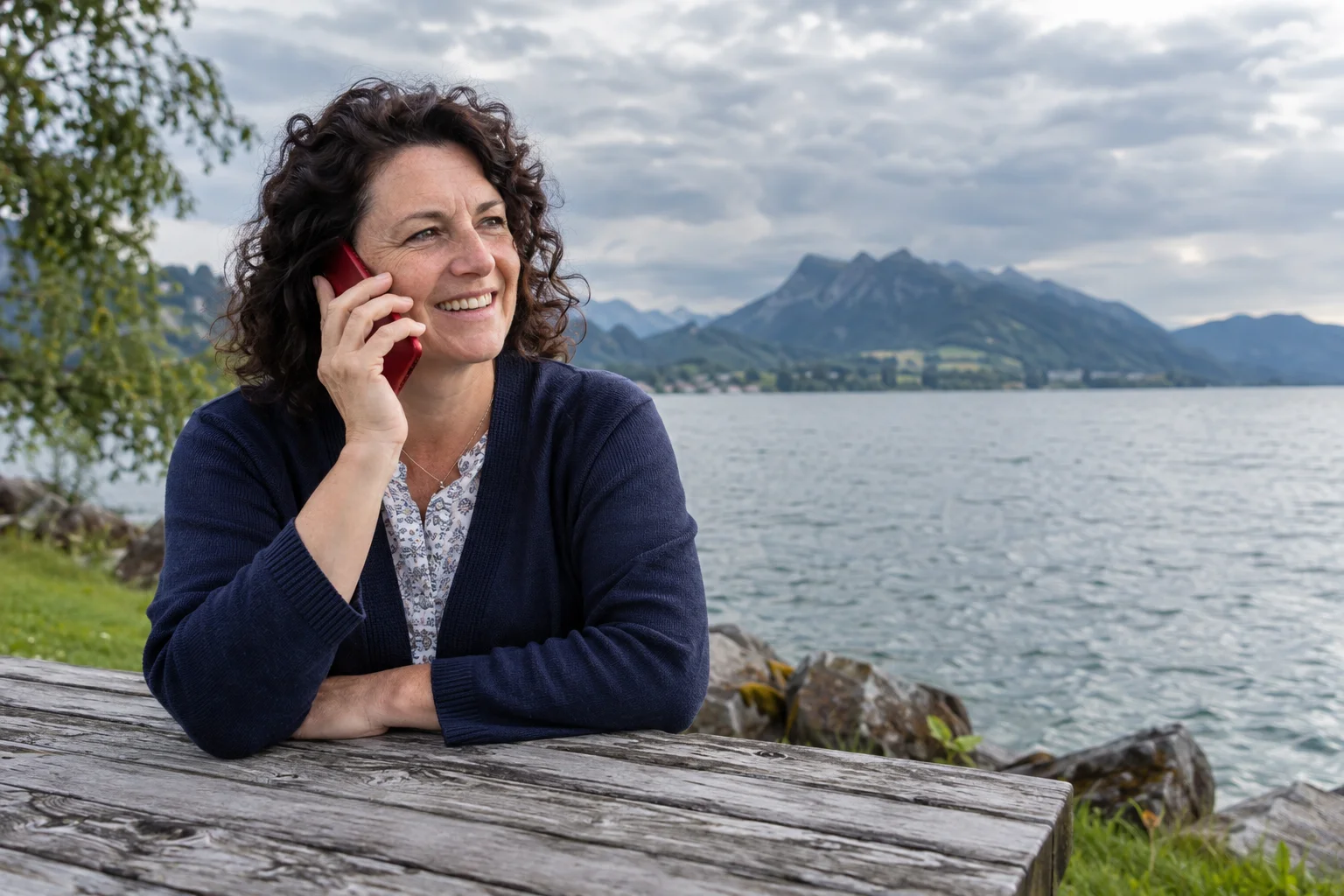 A woman talking on a red smartphone beside a lake with mountains beyond