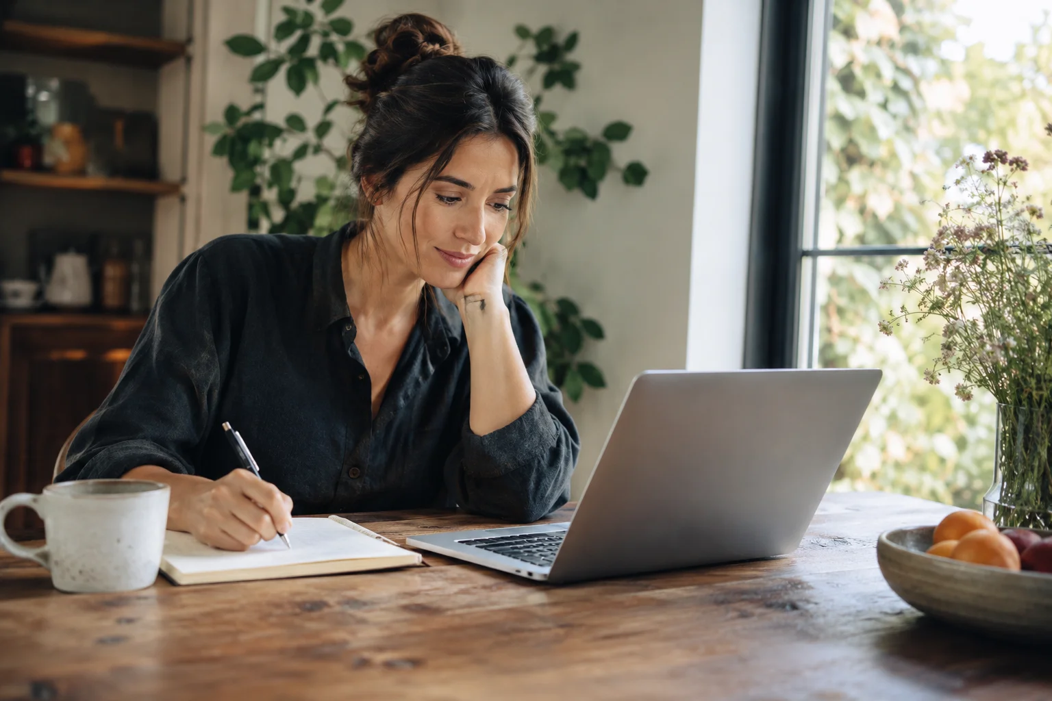 A woman writing notes beside an open laptop at a wooden table